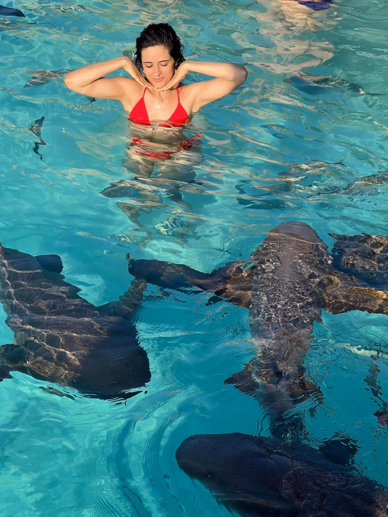 Close-up of Camila Cabello laughing while splashing in crystal clear ocean water wearing a red swimsuit.