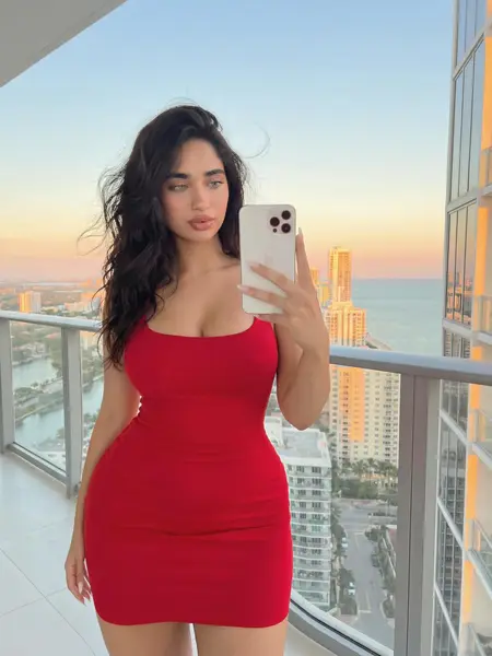 Woman in a red fitted dress taking a mirror selfie on a balcony at sunset overlooking the coastline