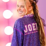 JoJo Siwa smiling on a pink carpet wearing a purple rhinestone jacket with "JOBI" branding