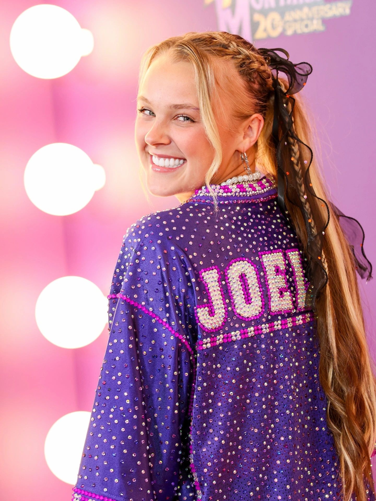 JoJo Siwa smiling on a pink carpet wearing a purple rhinestone jacket with "JOBI" branding