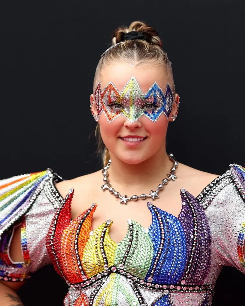 JoJo Siwa wearing a rainbow rhinestone face mask and matching crystal-embellished outfit