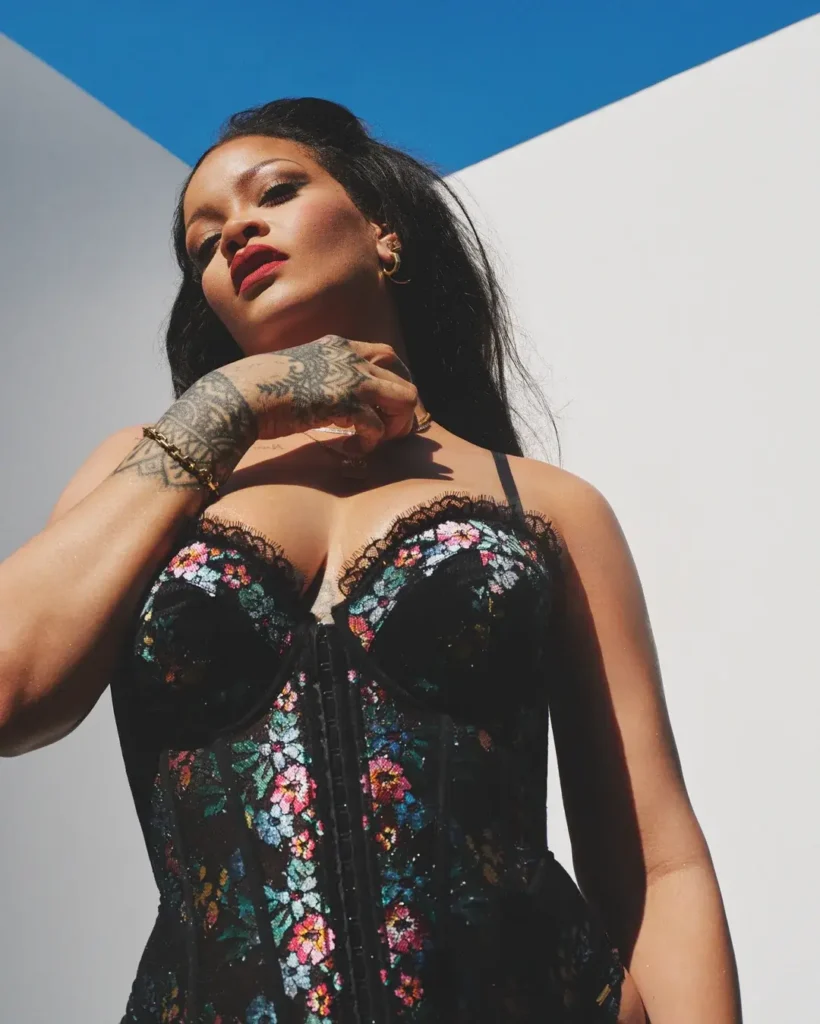Rihanna in a black floral lace-up corset bodysuit for Savage X Fenty, tilting her head back against a blue sky and white wall backdrop